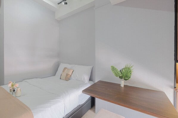 A bedroom with a bed, a nightstand, a small table with a plant, and a wall-mounted shelf. - Comfy And Well Studio At Tokyo Riverside Pik 2 Apartment (Teluknaga)