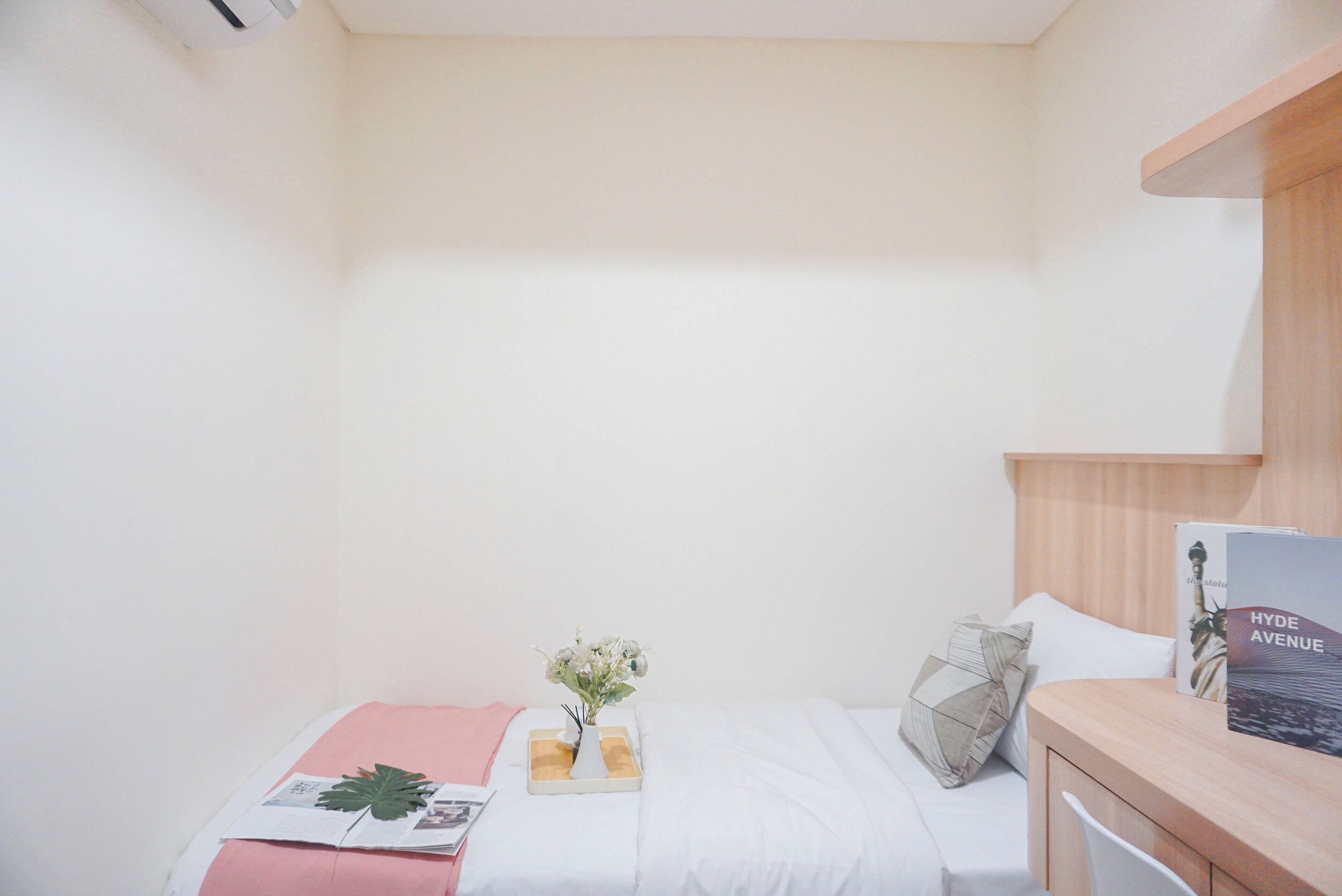 A small, neatly arranged bedroom with a single bed, a bedside table with a book, and a wall-mounted shelf.