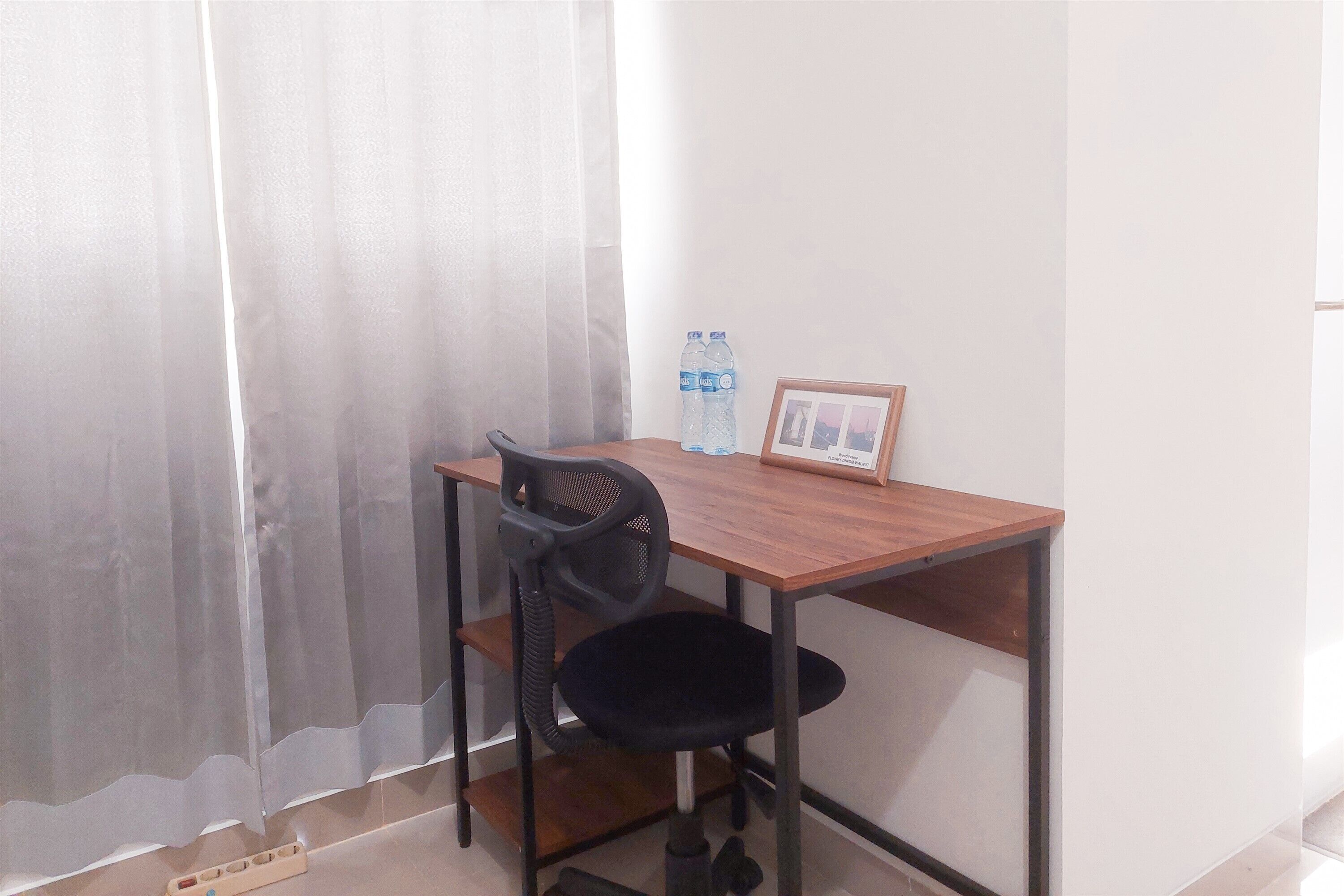 A small, neatly arranged room with a wooden desk, a black office chair, a water bottle, and a framed picture on the wall.