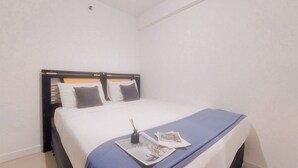 A neatly made bed with a blue bedspread and two pillows, a small table with a book and a bottle, and a plain white wall with a ceiling light.