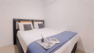A neatly made bed with a blue bedspread and two pillows, a small table with a book and a bottle, and a plain white wall with a ceiling light.