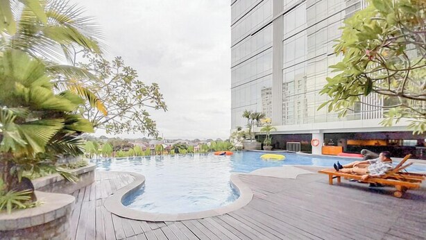 Una piscina en la azotea con cubierta de madera, rodeada de plantas tropicales, y un edificio moderno al fondo.