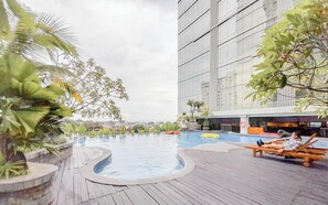 A rooftop pool with a wooden deck, surrounded by tropical plants, and a modern building in the background. - Cozy And Enjoy Living 1Br At Mataram City Apartment (Ngaglik)