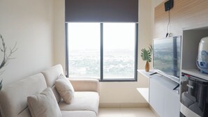 A modern living room with a sofa, a television, and a view of a cityscape through a large window. - Comfy Abode 2Br Apartment Pollux Chadstone (Cikarang)