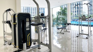 A gym with various exercise machines, a bench, and a view of a swimming pool through large windows. - Comfy Abode 2Br Apartment Pollux Chadstone (Cikarang)