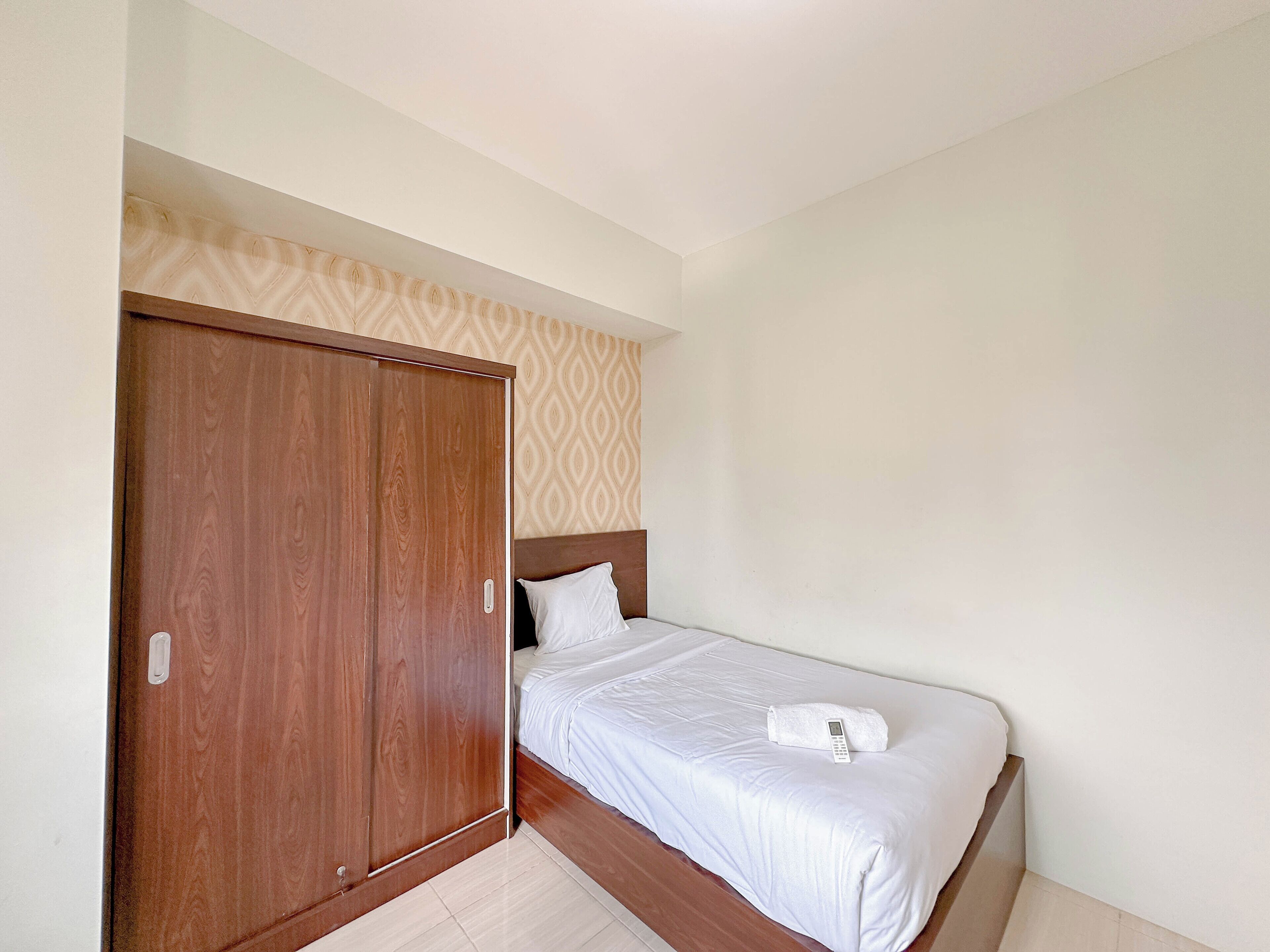 A bedroom with a wooden wardrobe, a bed with white bedding, and a patterned wallpaper above the bed.