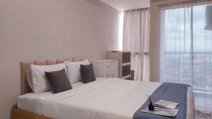 A neatly made bed with white linens and two pillows, a bedside table with a lamp, and a view of the city through a large window. - Good Studio Near Binus At Pacific Garden Apartment (Tangerang)