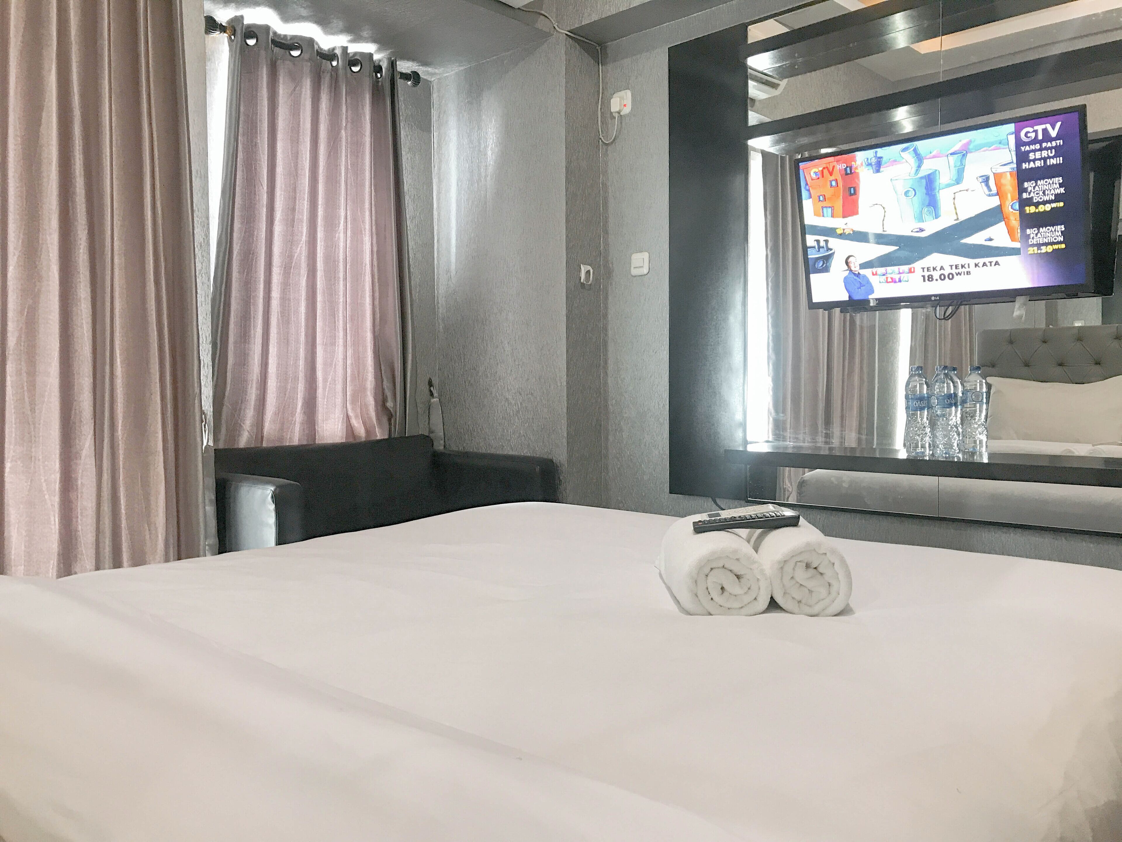 A hotel room with a bed, a TV mounted on the wall, a window with curtains, and a water bottle on the nightstand.