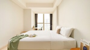 A hotel room with a large bed, a desk, and a chair. There is a window with curtains, and a view of buildings outside.