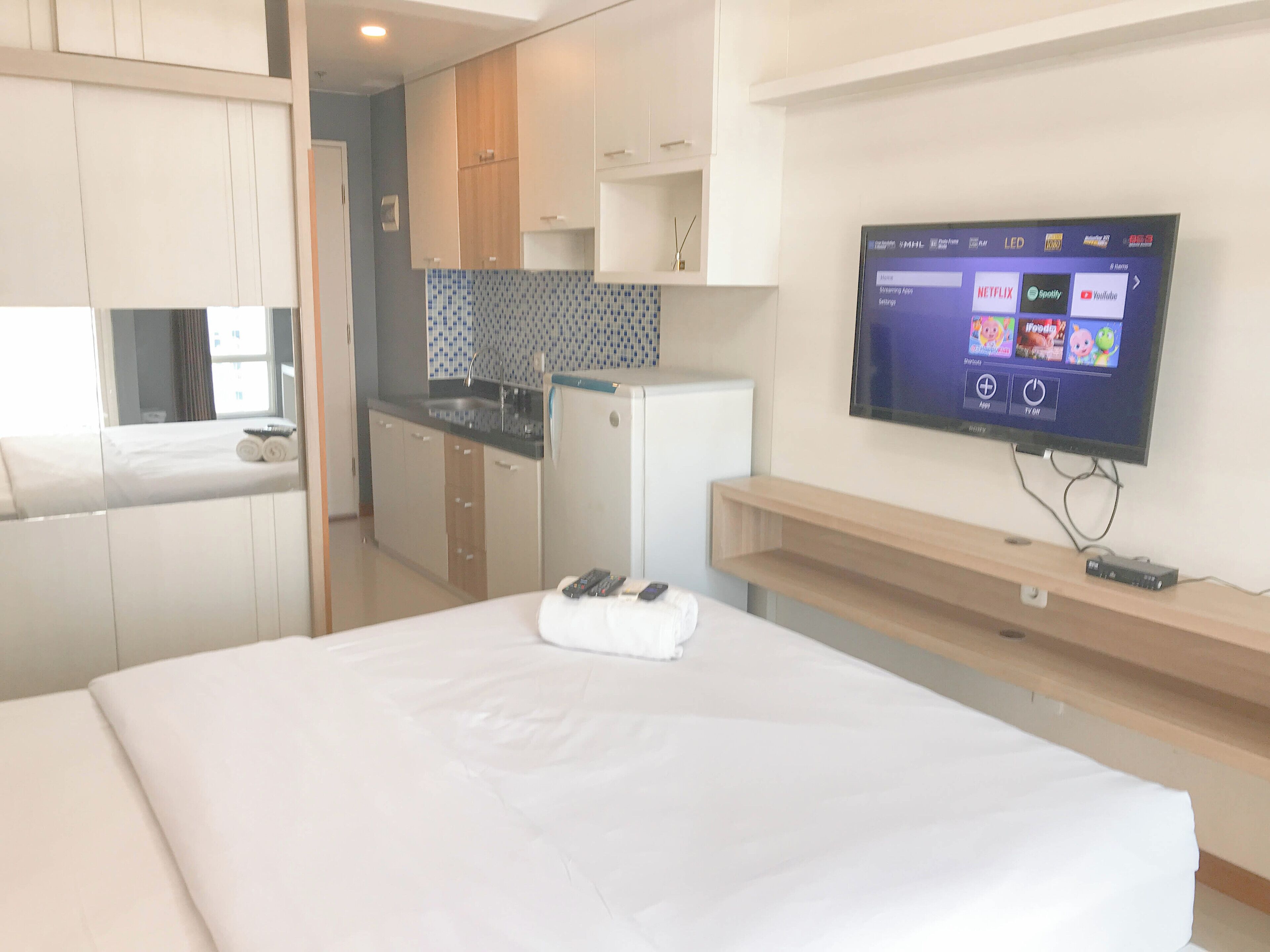 Ein Hotelzimmer mit Bett, einem an der Wand befestigten Fernseher, einer kleinen Küchenzeile und einem durchgehenden Türspalt erkennbaren Badezimmer.