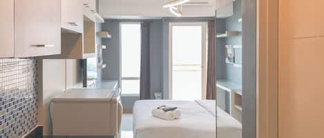 A modern hotel room with a bed, a small desk, a mirror, and a window with curtains.