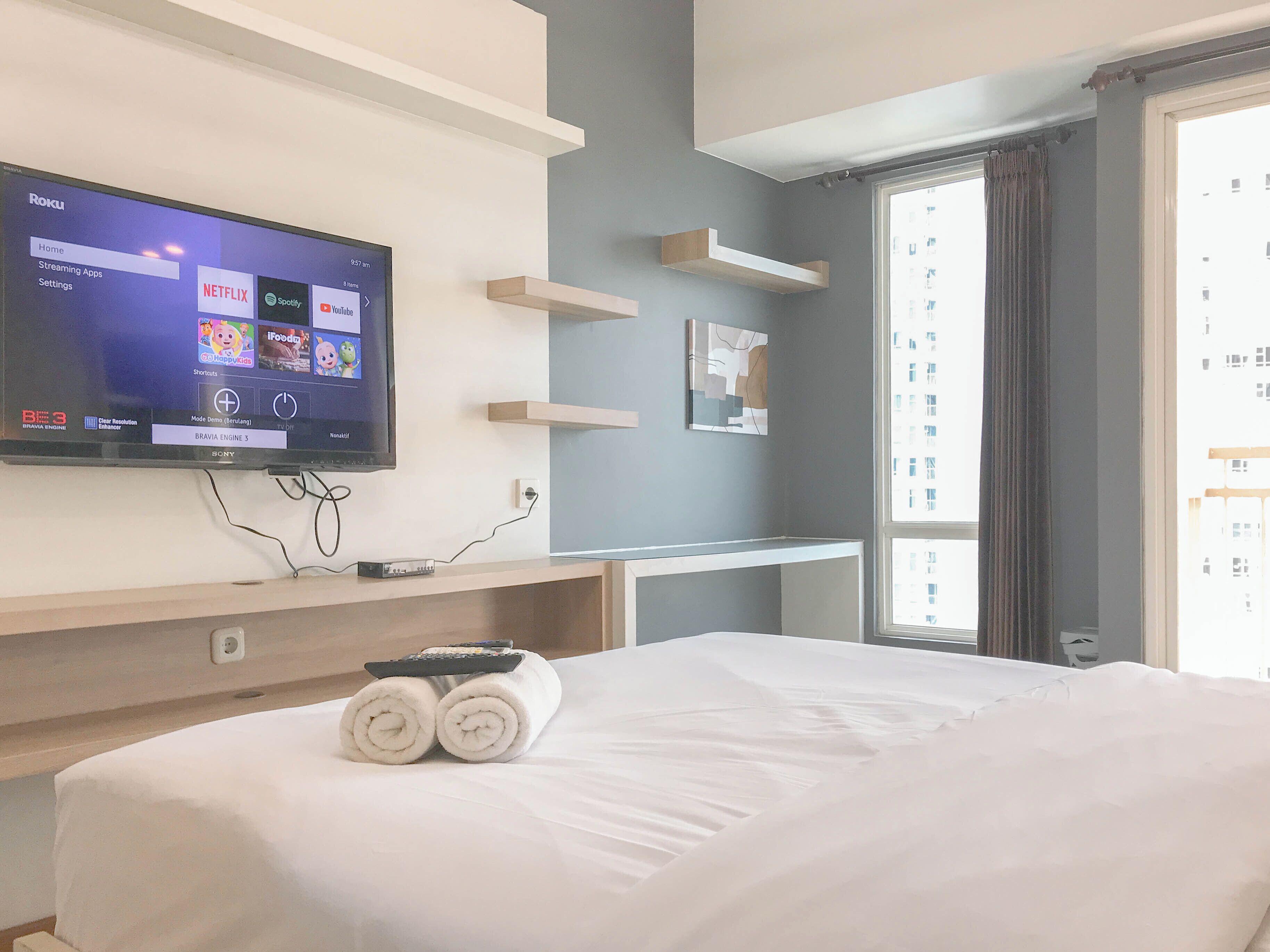 Ein Hotelzimmer mit einem Bett, einem an der Wand befestigten Fernseher, der Streaming-Apps anzeigt, einem Fenster mit Vorhängen und einem kleinen Tisch mit Fernbedienung und Handtüchern.