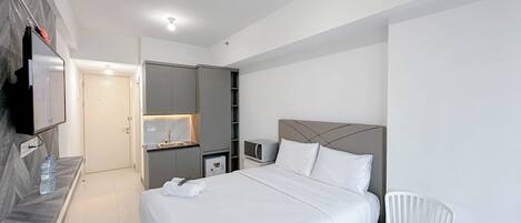 A modern hotel room with a large bed, a TV mounted on the wall, a small kitchenette, and a white chair.
