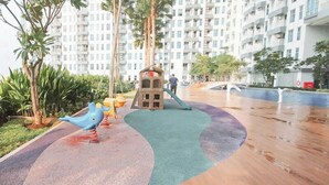 A playground with a slide and climbing structures, surrounded by palm trees and a residential building in the background. - Studio (No Kitchen) With City Light View Benson Supermall Mansion Apartment (Surabaya)
