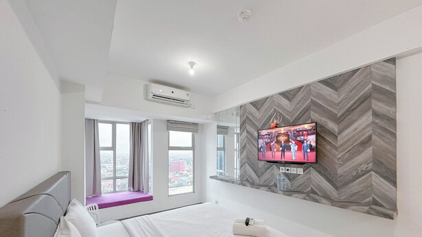 A hotel room with a bed, a TV mounted on the wall, a window seat, and a view of the city. - Studio (No Kitchen) With City Light View Benson Supermall Mansion Apartment (Surabaya)