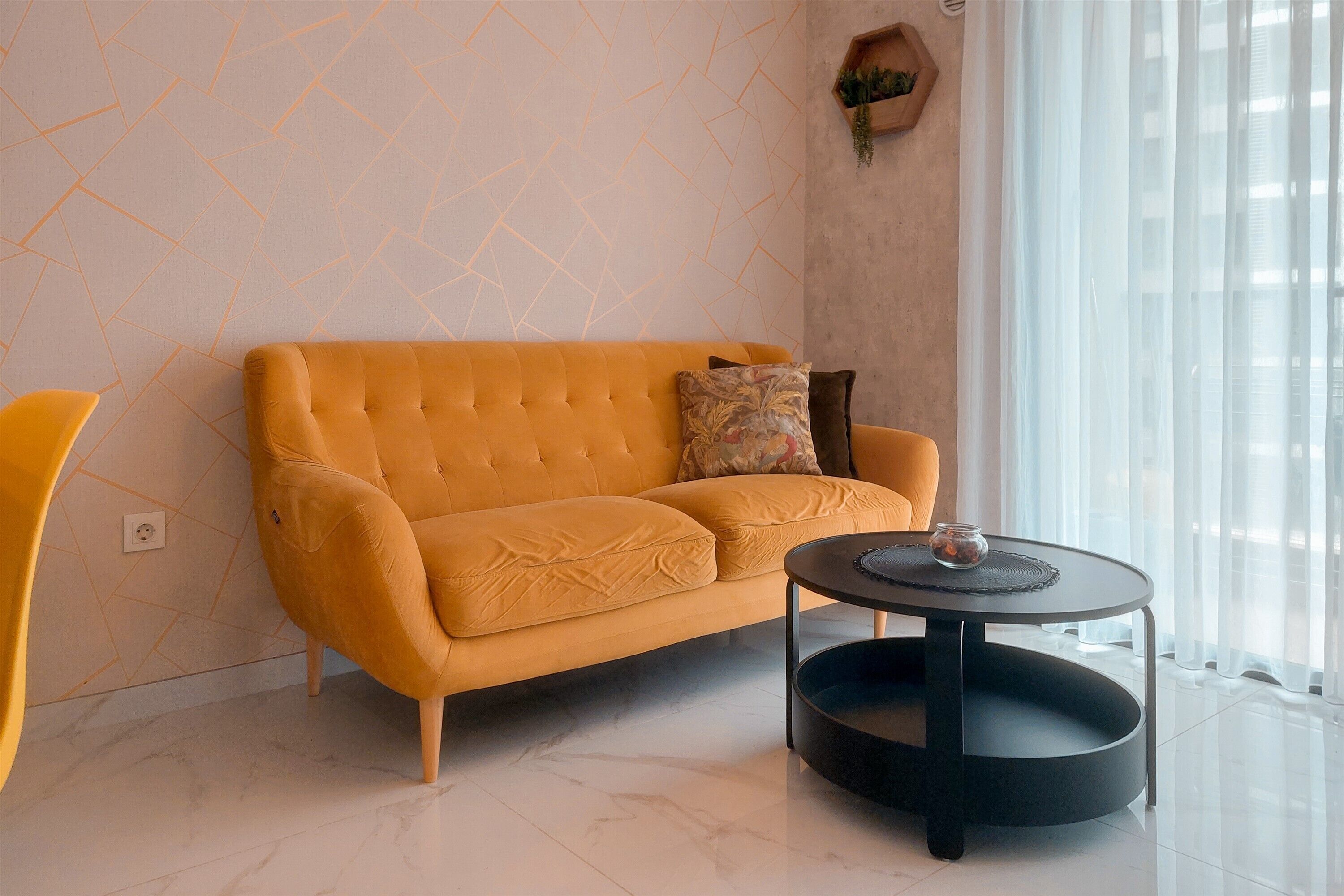 A modern living room with a yellow sofa, a round black coffee table, and a large window with sheer curtains.