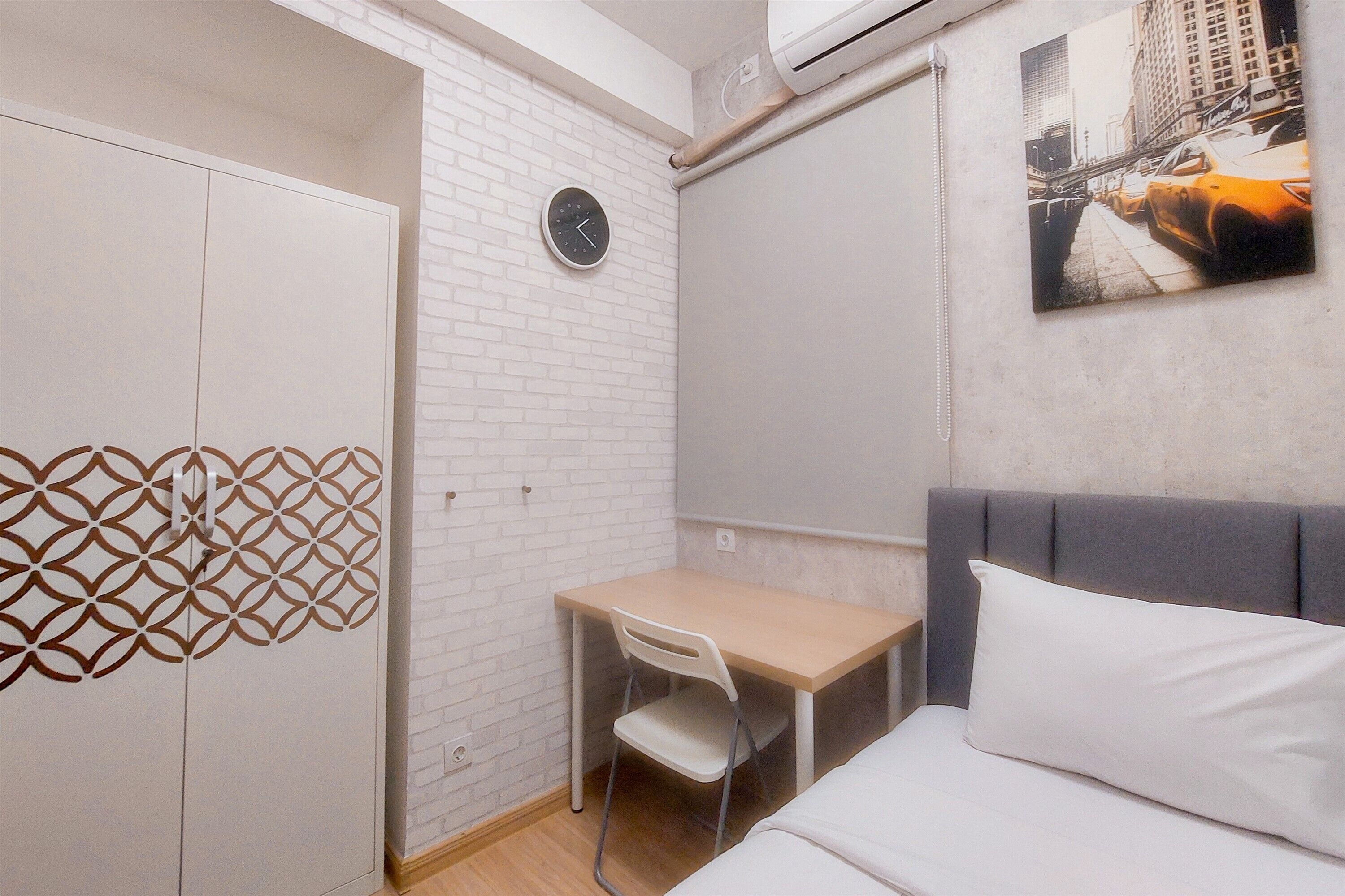 Una habitación pequeña y funcional con una cama, un escritorio y una silla. Hay un reloj en la pared y un cuadro con una vista urbana.