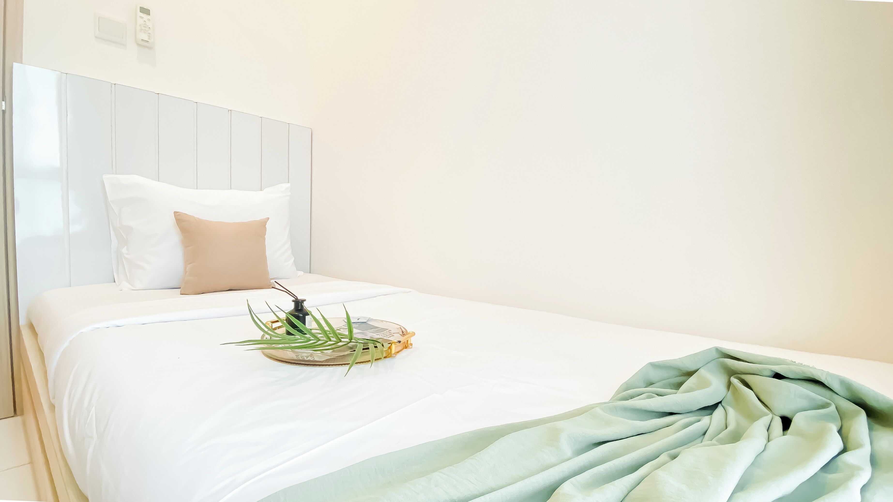A neatly made bed with white bedding, a beige pillow, a small plant, and a green throw blanket.