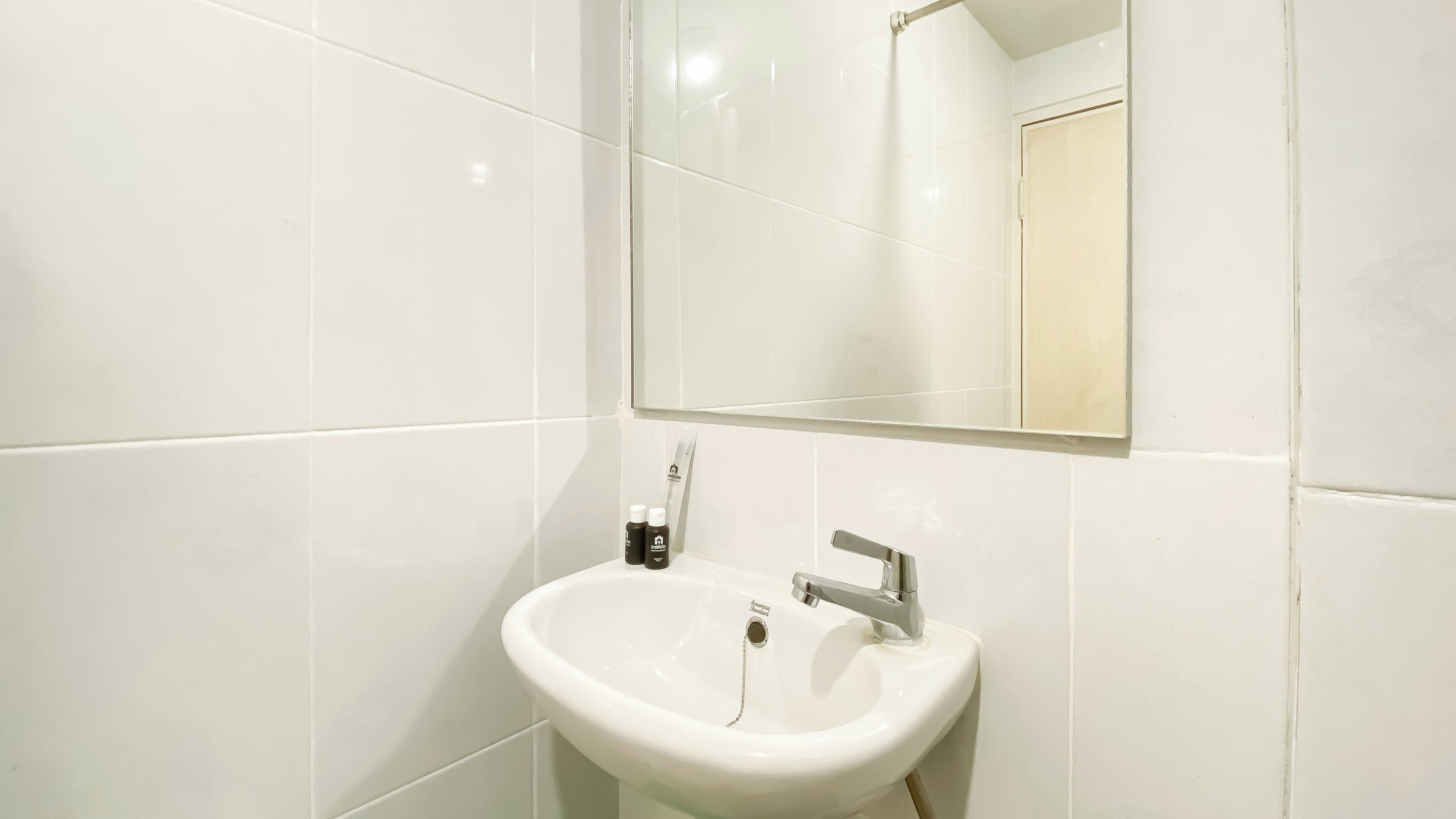 A bathroom with white tiles, a white sink, a mirror, and two bottles on the counter.