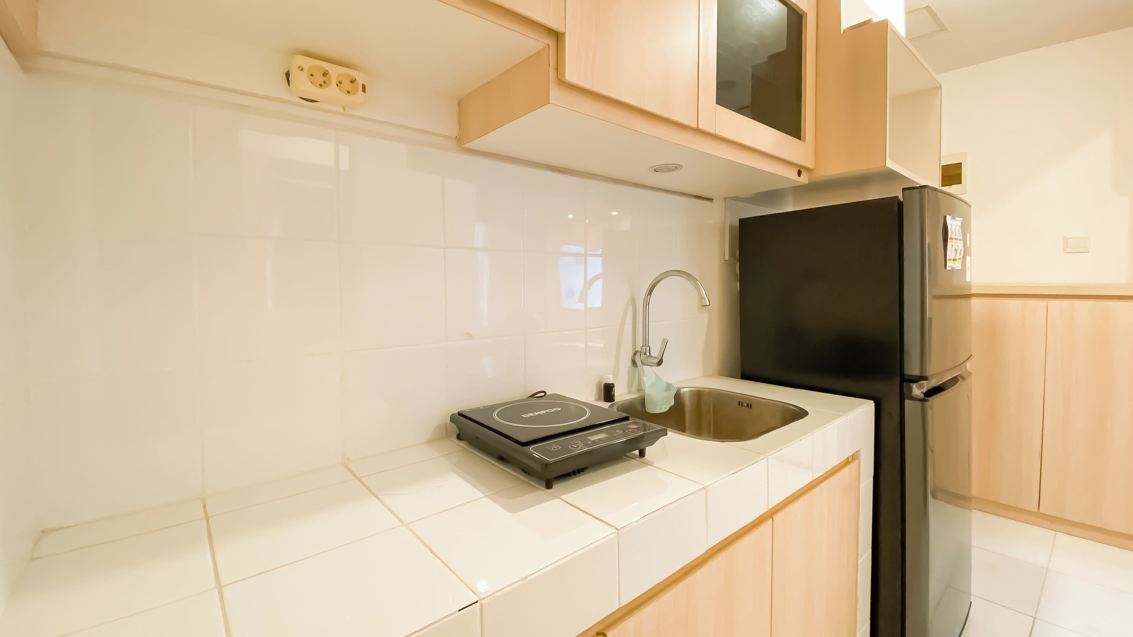 A modern kitchen with white tiles, a black induction cooktop, a stainless steel sink, and wooden cabinets.