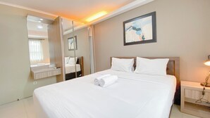 A neatly made bed with white linens and rolled towels, a bedside table with a lamp, a large mirror, and a painting on the wall. - The Modern Classic 2Br With Scenic View At Gateway Pasteur Apartment (Cimahi)