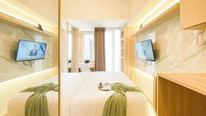 A hotel room with a large bed, two TVs mounted on the walls, a wooden vanity, and a bathroom visible through a glass door.