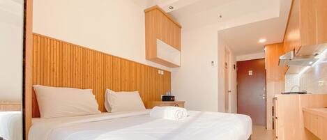 Habitación de hotel con cabecera de madera, cama con ropa blanca, mesita de noche pequeña y cocina integrada.