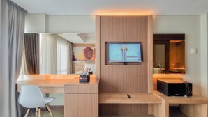 A modern hotel room with a flat-screen TV mounted on a wooden panel, a small desk with a chair, a microwave, and a large mirror. - Cozy Studio Apartment At Grand Mercure Yogyakarta (Yogyakarta)