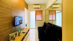 Een moderne woonkamer met een flatscreen televisie aan een houten wand, een donkergrijze bank, een houten salontafel met decoratieve objecten, en een grote spiegel die de kamer weerspiegelt.
