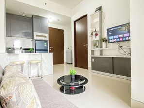 Una sala moderna con un sofá, una mesita redonda con una planta, una zona de cocina con un refrigerador y un televisor montado en la pared.