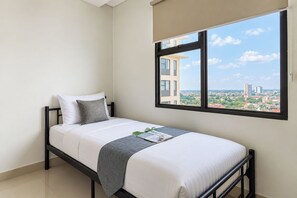 Una cama individual con cabecera, una ventana con vista a un paisaje urbano y una vista parcial de una silueta de la ciudad.