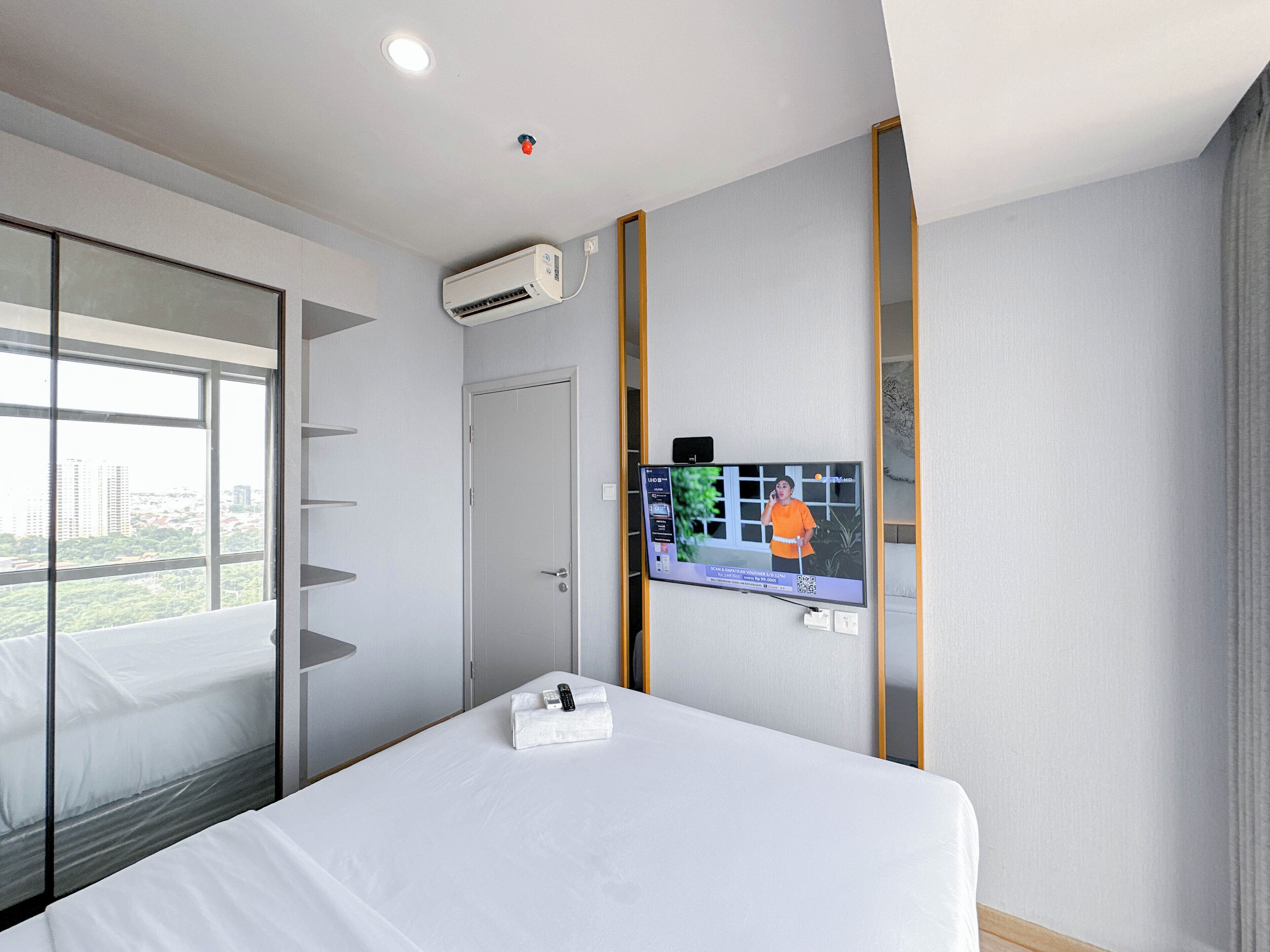 A hotel room with a bed, a TV on the wall, an air conditioning unit, and a window with a city view.
