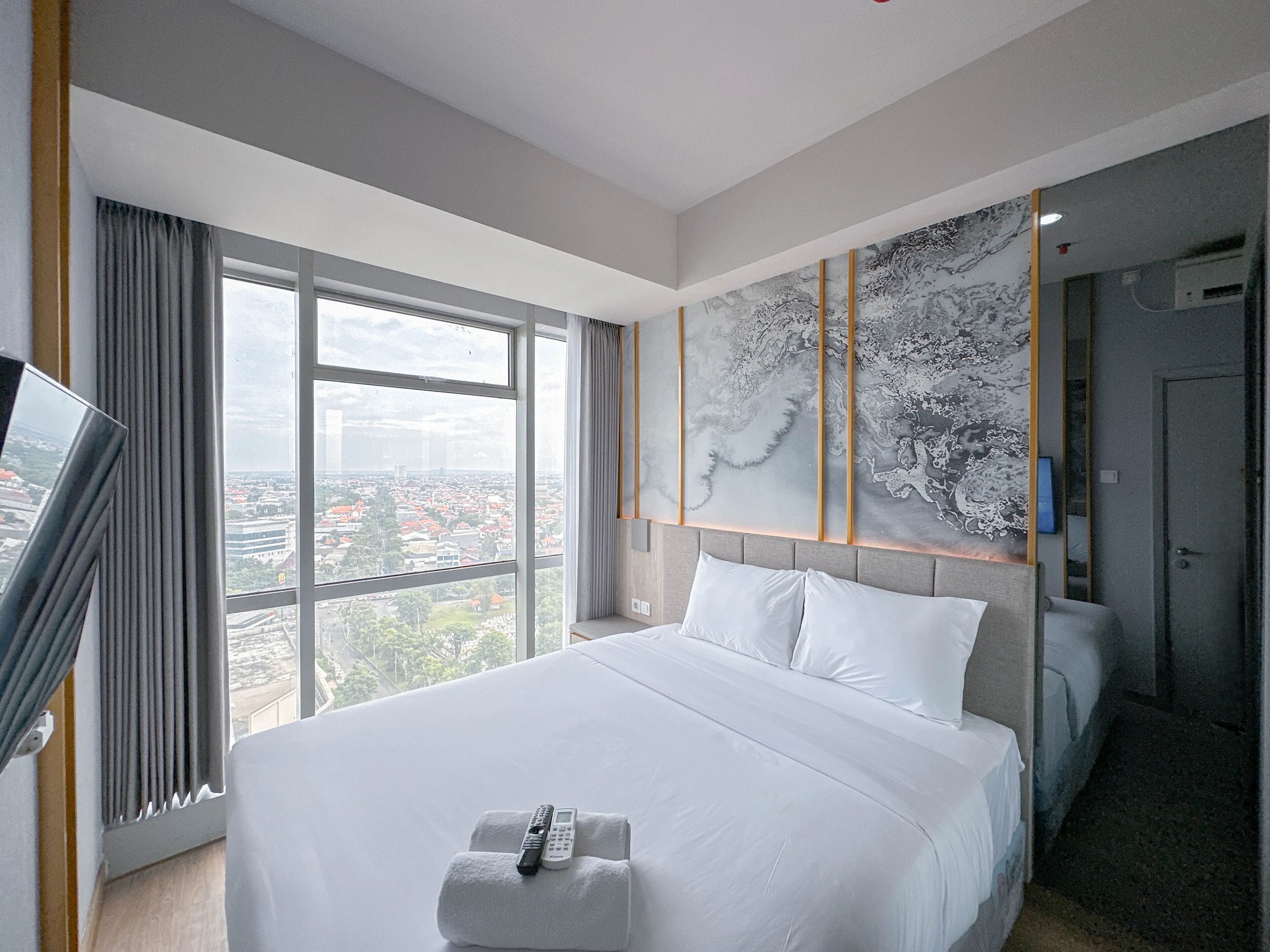 A hotel room with a large bed, a television, a view of the city, and a large abstract painting on the wall.