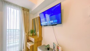 A modern living room with a flat-screen TV mounted on the wall, a wooden desk, a white sofa, and a large window with curtains. - Serene Studio At 25Th Floor Apartment Tokyo Riverside Pik 2 (Teluknaga)
