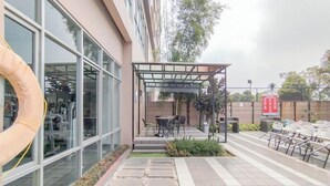 A building with large windows, a covered outdoor seating area with tables and chairs, and a pathway leading to the entrance. - The Chic 2Br Apartment At The Edge Bandung (Cimahi)