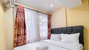 Una cama cuidadosamente tendida con sábanas blancas y una toalla enrollada, un cabecero tapizado en negro, y una ventana con cortinas de encaje blanco y cortinas con estampado floral.