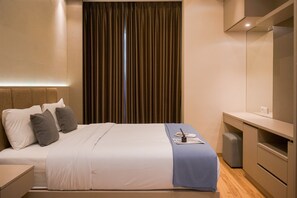 A modern hotel room with a neatly made bed, a small bedside table, a desk with a chair, and a large window with curtains. - Cozy 2Br Apartment M-Town Signature (Gading Serpong)