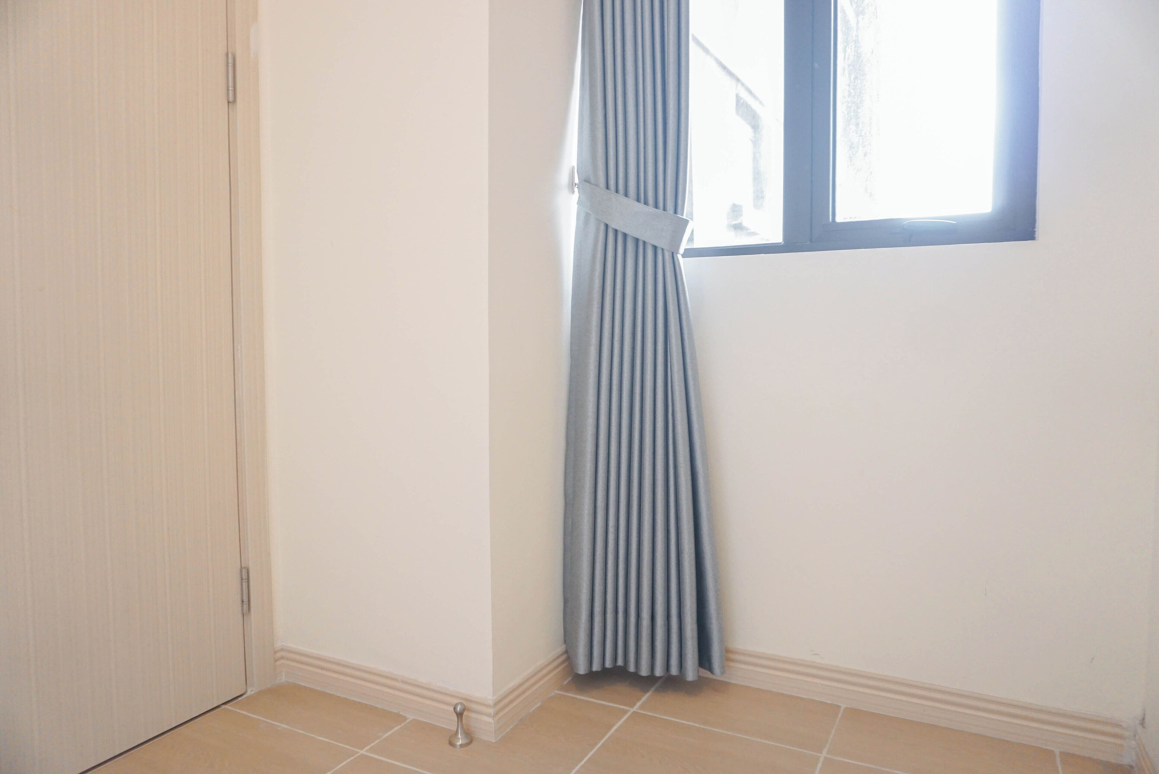 Una stanza con una porta chiusa, una finestra con tenda e pavimento piastrellato.