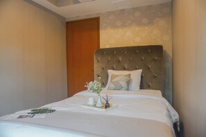 A neatly made bed with a tufted headboard, a bedside table with a vase of flowers, and a wooden door in the background. - Spacious And Luxury 3Br At Sudirman Tower Condominium Apartment (Jakarta)
