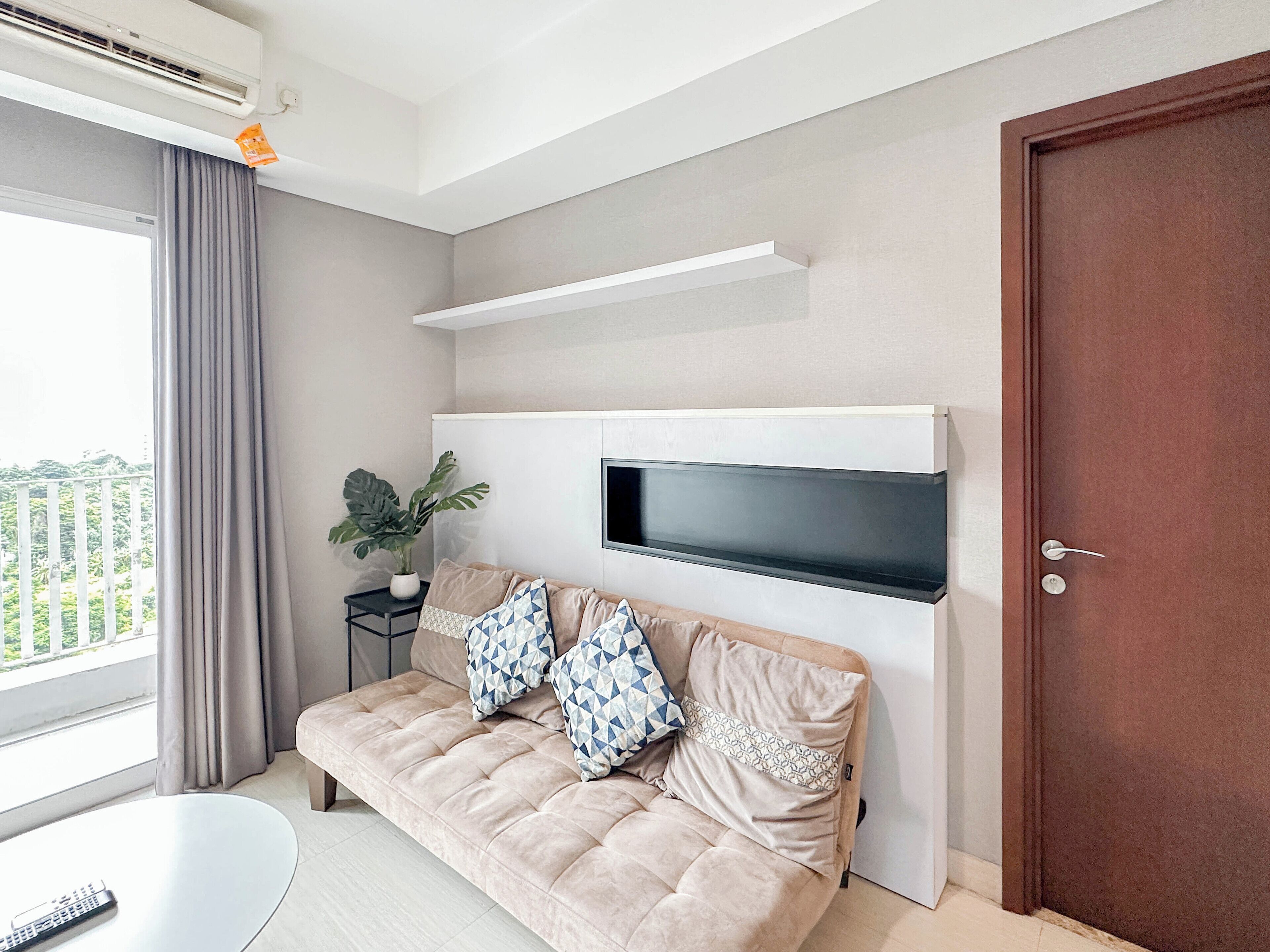Een moderne woonkamer met een beige bank, een flatscreen televisie aan een witte muur, een potplant en uitzicht op groen buiten.