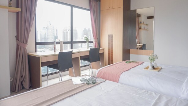 A modern hotel room with a large window, a desk with two chairs, a bed with a white comforter, and a mirror on the wall. - Best Location And Strategic 2Br At Tamansari Semanggi Apartment (Jakarta)