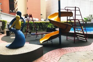 Un parque infantil con un tobogán amarillo y una estructura de escalada, rodeado de una zona pavimentada con una escultura decorativa.