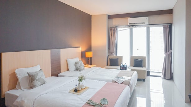 A hotel room with two beds, a sitting area with a chair, a window with curtains, and an air conditioning unit. - Comfortable Studio At Tamansari Semanggi Apartment (Jakarta)