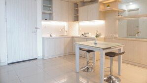 A modern kitchen with white cabinetry, a glass-top table, and two bar stools. - Comfy And Modern 2Br Grand Kamala Lagoon Apartment (Bekasi)