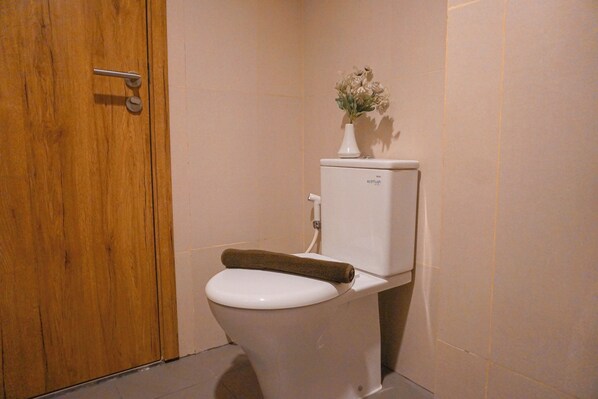 A toilet with a towel on the seat, a white vase with flowers on the tank, and a wooden door. - Cozy Studio Apartment At LRT City Jatibening (Bekasi)