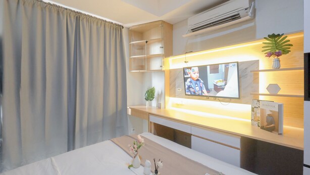 A hotel room with a bed, a TV mounted on the wall, a shelf with a plant, and a small table with a vase. - Homey And Comfortable Studio Apartment At Daan Mogot City (Jakarta)