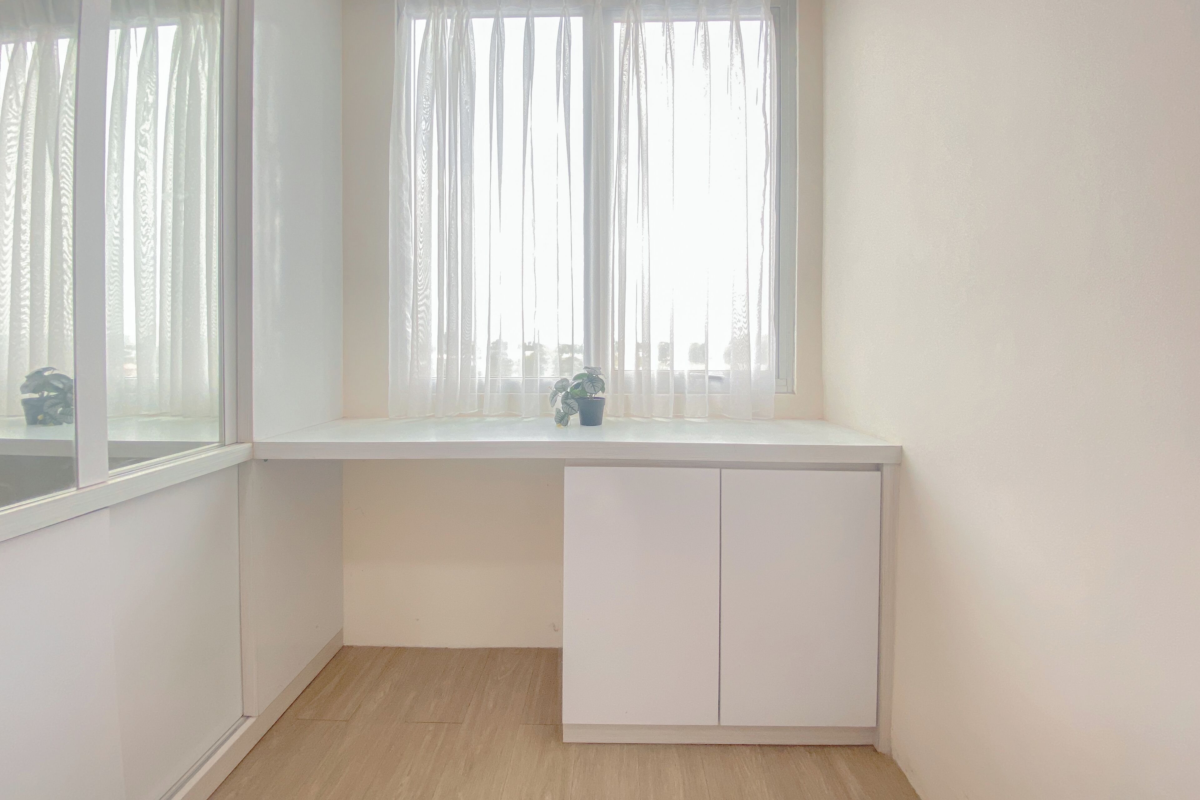 Une pièce d’angle avec un bureau blanc, une fenêtre ornée de rideaux légers et une petite plante posée sur le bureau.