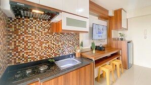A modern kitchen with a checkered backsplash, wooden cabinets, a black countertop, a built-in stove, a sink, and a wooden dining area with stools. - Comfort Stay 1Br With Extra Room At Bassura City Apartment (Jakarta)