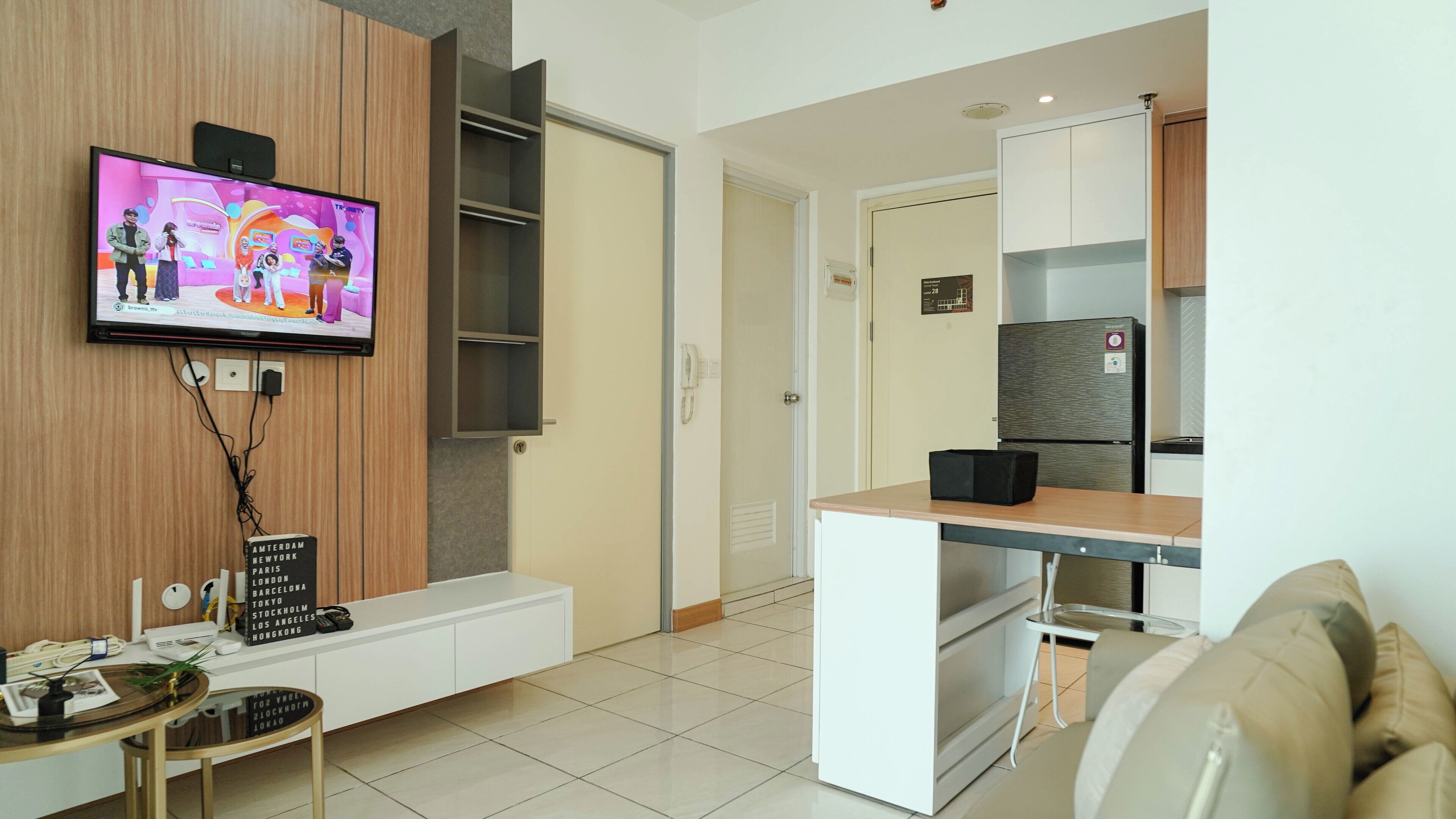 Un soggiorno moderno con una TV a schermo piatto montata a parete, una parete d'accento in legno, un divano bianco, un tavolino con una pianta, e un angolo cottura con frigorifero e pensili.