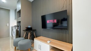 A modern living room with a flat-screen TV displaying 'NETFLIX', a washing machine, a small table with two bottles, and a wooden bench. - Brand New And Homey Studio At Gold Coast Apartment Pik (Jakarta)